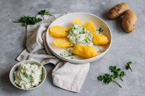 Pellkartoffeln mit Avocado-Kräuterquark in weißem Teller neben Kartoffeln und Kräutern vor hellem Hintergrund.