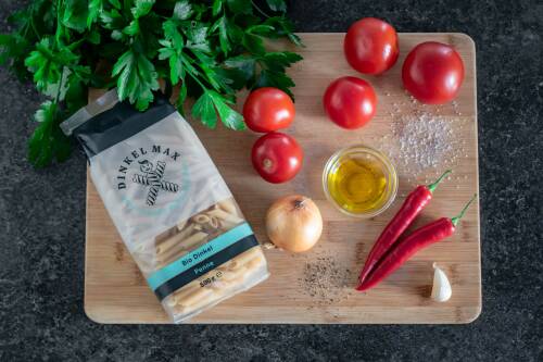 Zutaten für Penne all'Arrabbiata auf einem Holzbrett: Petersilie, Tomaten, Zwiebel, Knoblauch, Olivenöl, Penne, Pfeffer, Salz, Chili-Schoten