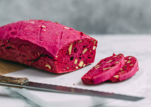 Knallig pinkes Nussbrot, angeschnitten auf einem Brett, daneben ein Messer. Von der Seite fotografiert.