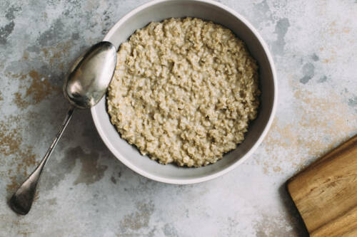 Porridge in weißer Schüssel neben Löffel und Schneidebrett vor hellem Hintergrund.