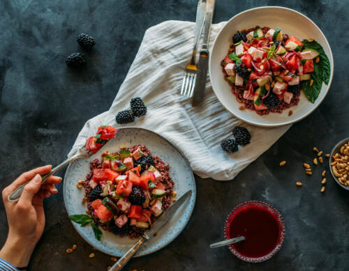 Quinoasalat auf zwei Tellern mit Beeren und Besteck in Hand vor dunklem Hintergrund.