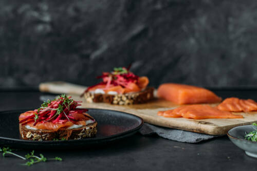 Räucherlachs und Rote Bete auf Teller und Schneidebrett mit Vollkornbrot vor dunklem Hintergrund.