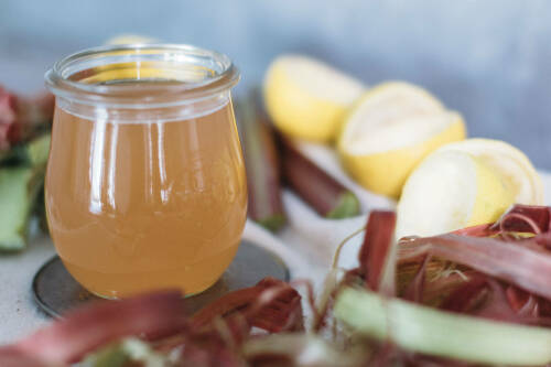 Rhabarber-Sirup in Glas neben Rhabarber vor hellem Hintergrund.