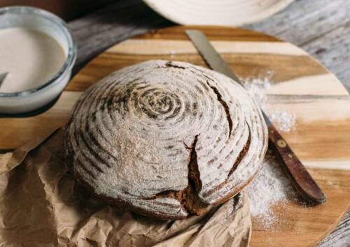 Sauerteig-Roggenbrot auf Schneidebrett neben Messer und Mehl vor hellem Hintergrund.