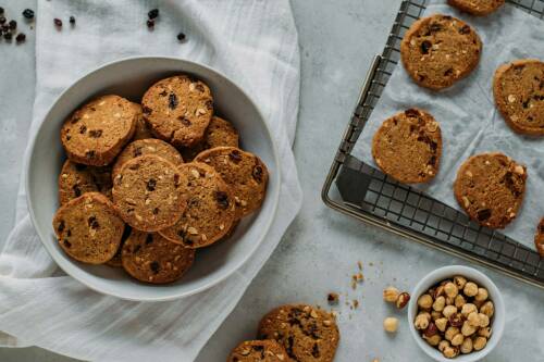 Rosinen-Haferflocken-Kekse in Schüssel und auf Backbleck vor hellem Hintergrund.