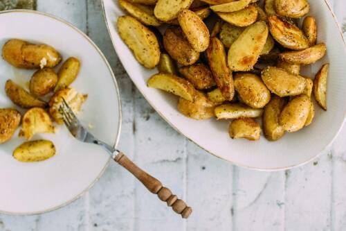Rosmarinkartoffeln vom Blech mit Gabel auf weißen Schüsseln und Teller vor hellem Hintergrund.