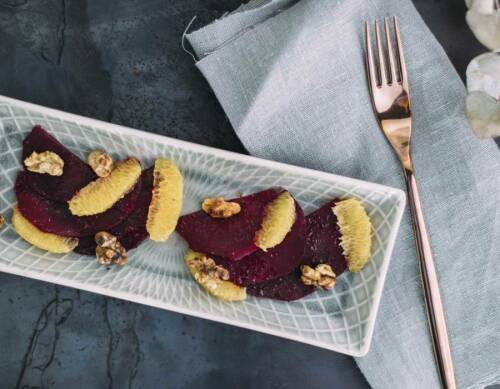 Rote Bete Carpaccio mit Orangen und Walnüssen auf Teller neben Gabel vor dunklem Hintergrund.