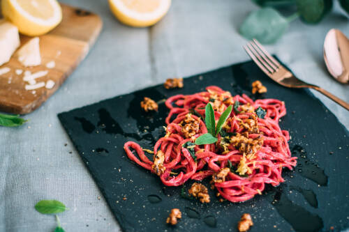 Schwarze Schieferplatte mit pinken Rote Bete Nudeln und Salbei-Zitronen-Butter mit karamellisierten Walnüssen darauf. Im Hintergrund ein Schneidebrett mit zwei halben Zitronen, von oben fotografiert.