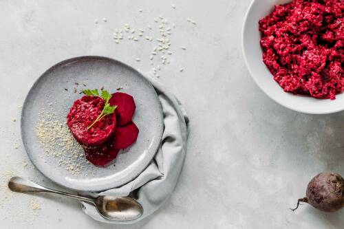 Rote-Bete-Risotto auf Teller und Schüssel neben Löffel vor hellem Hintergrund.