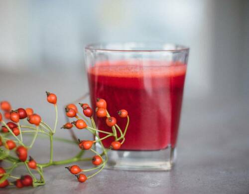 Roter Apfel-Karottensaft in Glas neben Beeren vor hellem Hintergrund.