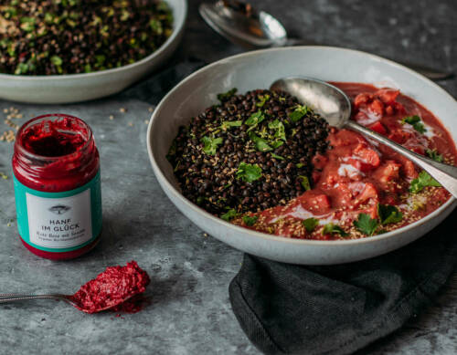 Rotes Blumenkohl-Curry mit Belugalinsen in weißer Schüssel neben Glas Rote Bete Paste vor grauem Hnitergrund.