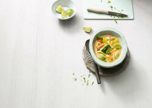 Rotes Gemüsecurry in einer weißen Schüssel, daneben ein Schneidebrett und eine Schüssel voll mit Limetten. Von oben fotografiert.