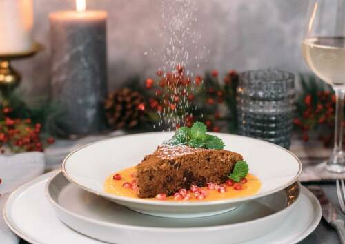 Weihnachtlicher Rotweinkuchen auf Mangospiegel dazu Granatapfelkerne. Im Hintergrund festliche Dekorationen, Kerze und Zweige. Von der Seite fotografiert.