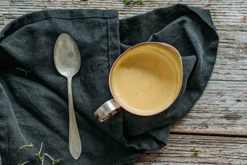 Sauce Hollandaise in Kännchen neben Löffel vor dunklem Hintergrund.