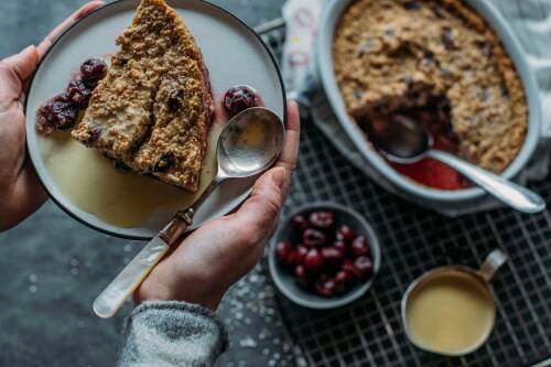 Sauerkirschauflauf mit gepufftem Amaranth auf Teller und in Auflaufform mit Händen vor hellem Hintergrund.