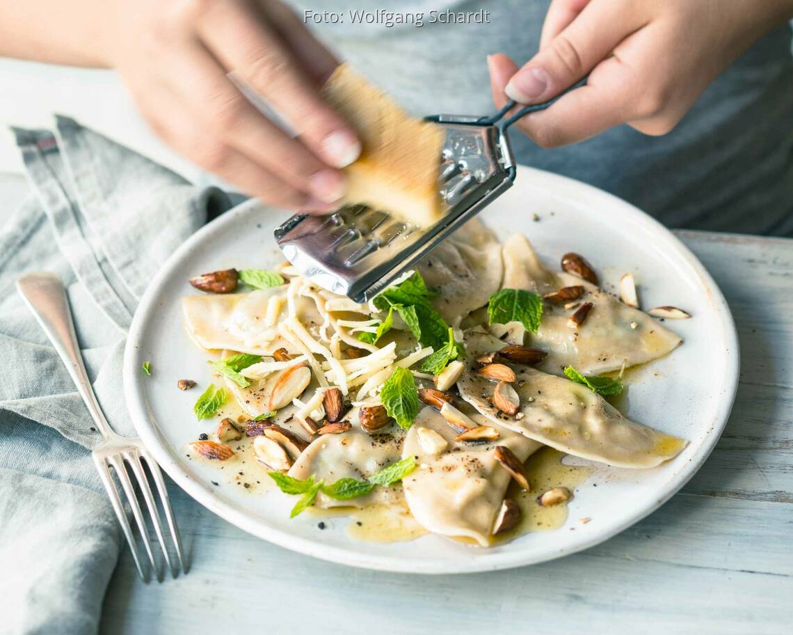 Selbstgemachte Schlutzkrapfen mit Pak Choi gefüllt auf weißem Teller, Holztisch. Frau reibt Parmesan darüber.
