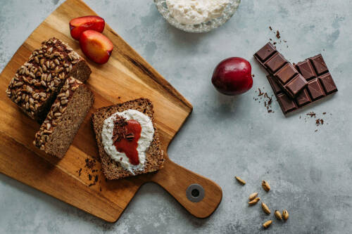 Schnelles Frühstücksbrot mit Ricotta und Pflaumenmus neben Pflaumen auf Schneidebrett vor hellem Hintergrund.