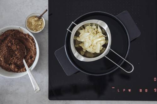 Kakaobutter in Topf neben Kakao auf Herd vor dunklem Hintergrund.