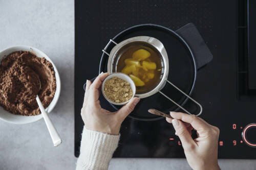 Geschmolzene Butter in Topf mit Hand und Zucker neben Kakao vor dunklem Hintergrund.