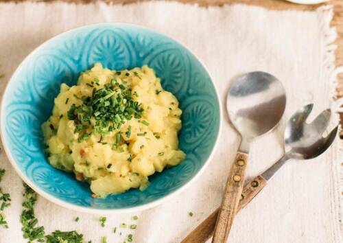 Portion schwäbischer Kartoffelsalat in einer blauen Schüssel, daneben Salatbesteck. Von oben fotografiert.