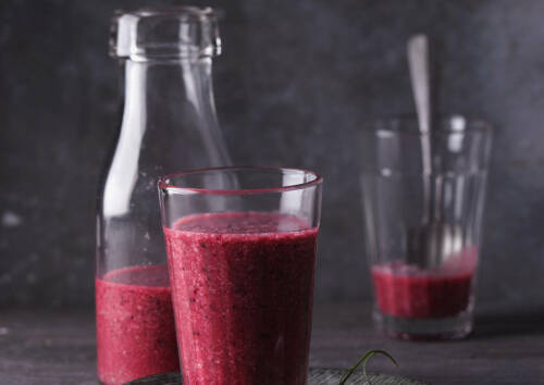 Eine Flasche und zwei Gläser mit einen dunkelroten Smoothie aus schwarzer Johannisbeere gefüllt. Hintergrund ist dunkelgrau, von der Seite fotografiert.