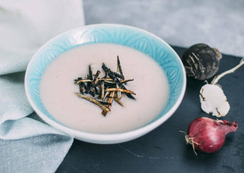 Rosa Schwarzrettichsuppe mit Gemüsechips in einer blauen Schüssel, daneben Zwiebel und Rettich-Knolle, von oben fotografiert.