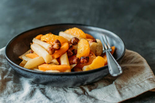 Schwarzwurzel mit Kaki und Orange auf schwarzem Teller vor dunklem Hintergrund.