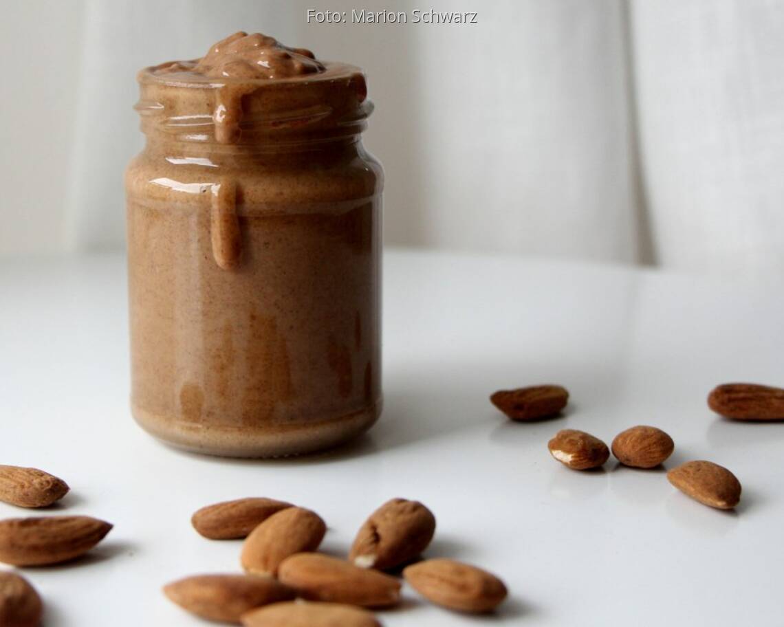 Mandelmus selbstgemacht in Schraubglas mit ungeschälten Mandeln als Deko auf weißem Tisch