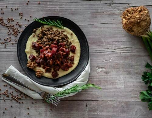 Selleriepüree mit Rote-Bete-Ragout und Rosmarinlinsen in schwarzem Teller neben Besteck und Rosmarinzweigen auf Holztisch.