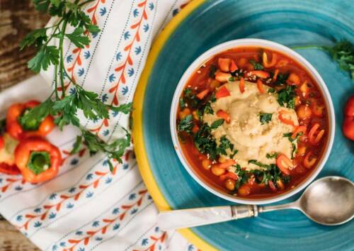 Knallig rotes Shakshuka in der veganen Variante in einer Schüssel auf einem blauen Teller. daneben Petersilie und Paprikaschoten. Von oben fotografiert.