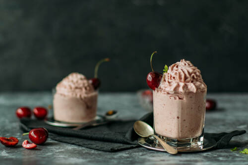 Softeis mit Kirschen in Gläsern vor dunklem Hintergrund neben Kirschen und Löffeln.