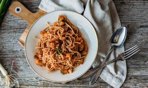 Spaghetti Verdure auf weißem Teller und Schneidebrett neben Besteck vor hellem Hintergrund.