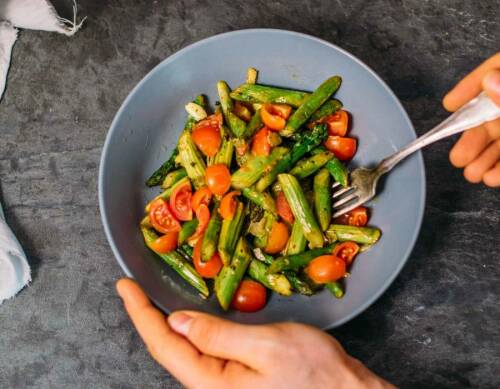 Spargel-Tomaten-Salat in Teller mit Händen vor dunklem Hintergrund.