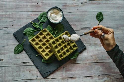 Spinat-Buchweizen-Waffeln auf Schiefertafel mit Hand und Schmand vor hellem Hintergrund.
