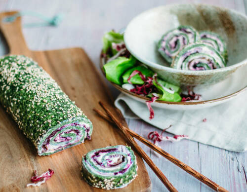 Spinat-Roulade aufgeschnitten neben Stäbchen auf Schneidebrett vor weißem Hintergrund.