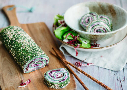 Spinat-Roulade mit Frischkäse und Radicchio auf einem Holzbrett, angeschnitten daneben Stäbchen, von oben fotografiert.