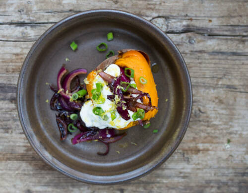 Süßkartoffeln mit Zitronenschmand und gerösteten Zwiebeln auf dunklem Teller vor hellem Hintergrund.