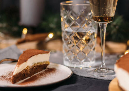 Italienische Tiramisu-Torte mit Kakao bestäubt auf einem Teller. Im Hintergrund ein festlich gedeckter Tisch, von vorne fotografiert.