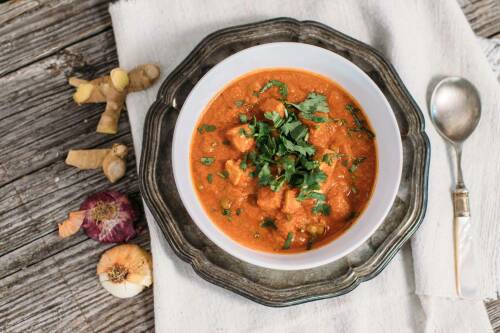 Tofu Masala in Suppenteller neben Ingwer und Löffel vor hellem Hintergrund.