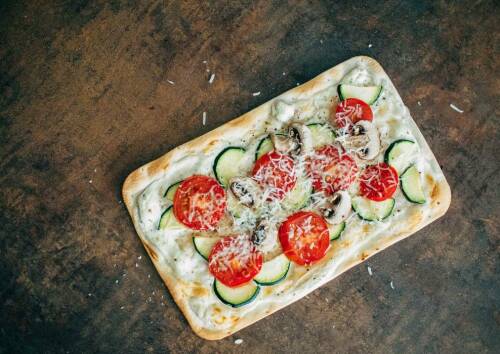 Flammkuchen mit Tomaten, Zucchini und Pilzen von oben fotografiert vor dunklem Hintergrund.
