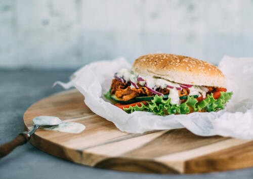 Burger mit Jackfruit auf einem Holzbrett mit Backpapier vor grauem Hintergrund, von der Seite fotografiert.