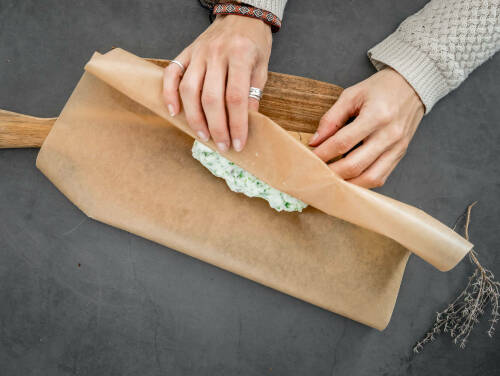 Vegane Kräuterbutter eingerollt in Backpapier mit Händen vor dunklem Hintergrund.