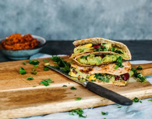 Vegane Quesadillas aufgeschnitten auf Schneidebrett neben Messer und Kidneybohnenpaste vor dunklem Hintergrund.