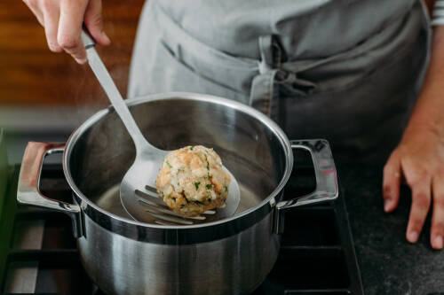 Semmelknödel in kochendem Wasser mit Schöpflöffel und Händen vor hellem Hintergrund.