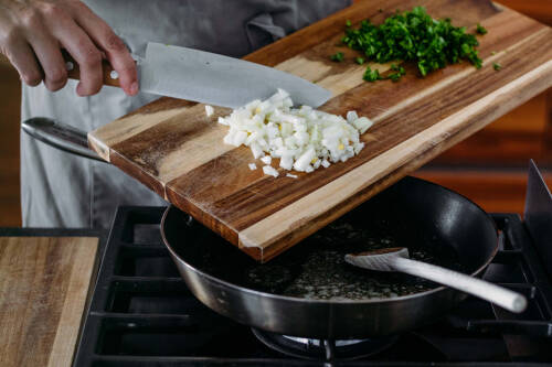 Gehackte Zwiebeln und Petersilie auf Schneidebrett über Pfanne vor hellem Hintergrund.