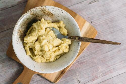 Veganer Kartoffelbrei in weißer Schüssel auf Schneidebrett mit Löffel.