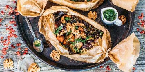 In Backpapier gegartes Gemüse auf einem dunklen Tablett. Daneben Kräuterdressing, Walnüsse und ein Löffel, von oben fotografiert.