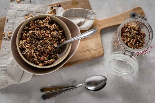 Veganes Müsli ohne Zucker in weißer Schüssel auf Schneidebrett neben Weckglas und Löffeln vor hellem Hintergrund.