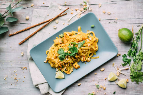 Veganes Pad Thai auf Teller neben Kräutern und Stäbchen vor hellem Hintergrund.