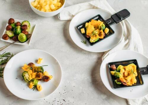 Veganes Raclette-Pfännchen mit Rosenkohl und Maronen auf weißen Tellern angerichtet.